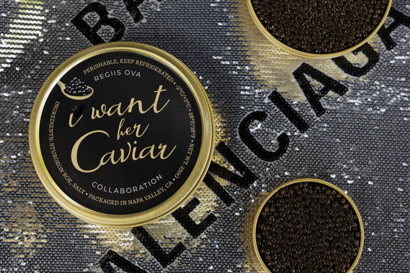 An I Want Her Caviar tin, a Regiis Ova collaboration, beside two open tins of caviar on silver sequins