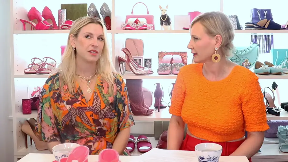 Two women in floral and orange tops talking on the I Want Her Shoes podcast set, in front of shelves of designer heels — the Samantha Brown conversation on style as strategy
