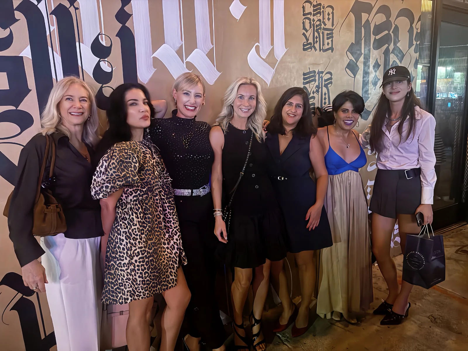 Seven I Want Her Shoes Club women smiling together in evening outfits, from leopard print to a blue satin top, in front of a black-and-gold calligraphy mural